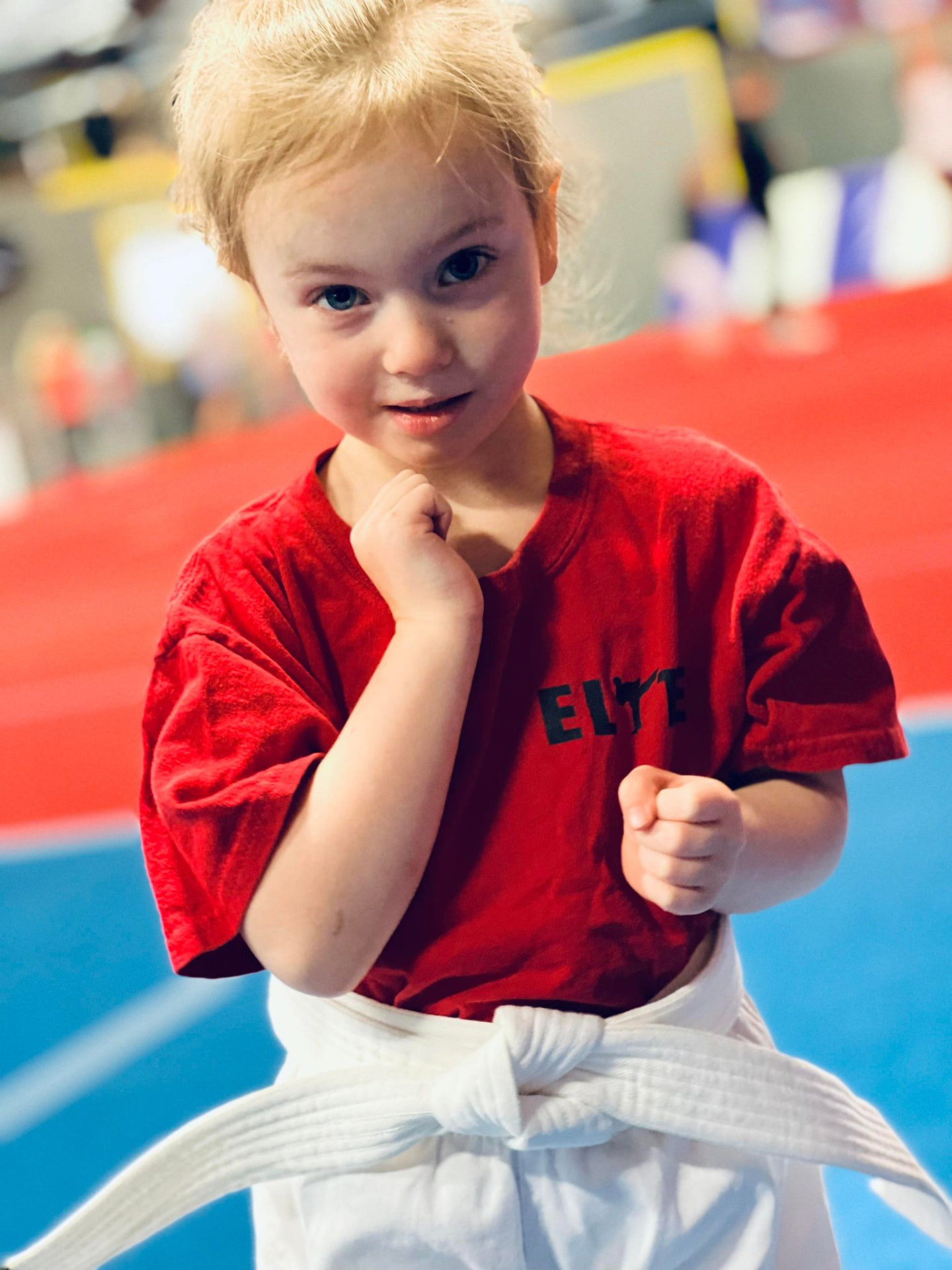 A young Mini Elite athlete in a red Elite shirt and white belt with hands raised, ready to train.