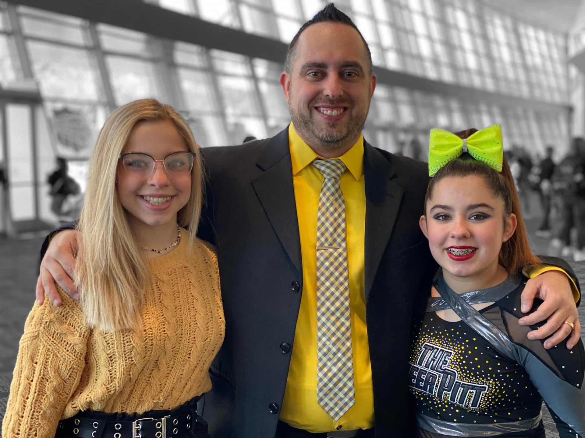 A man and two young athletes, one in her Cheer Pitt uniform, posing together at a cheer competition