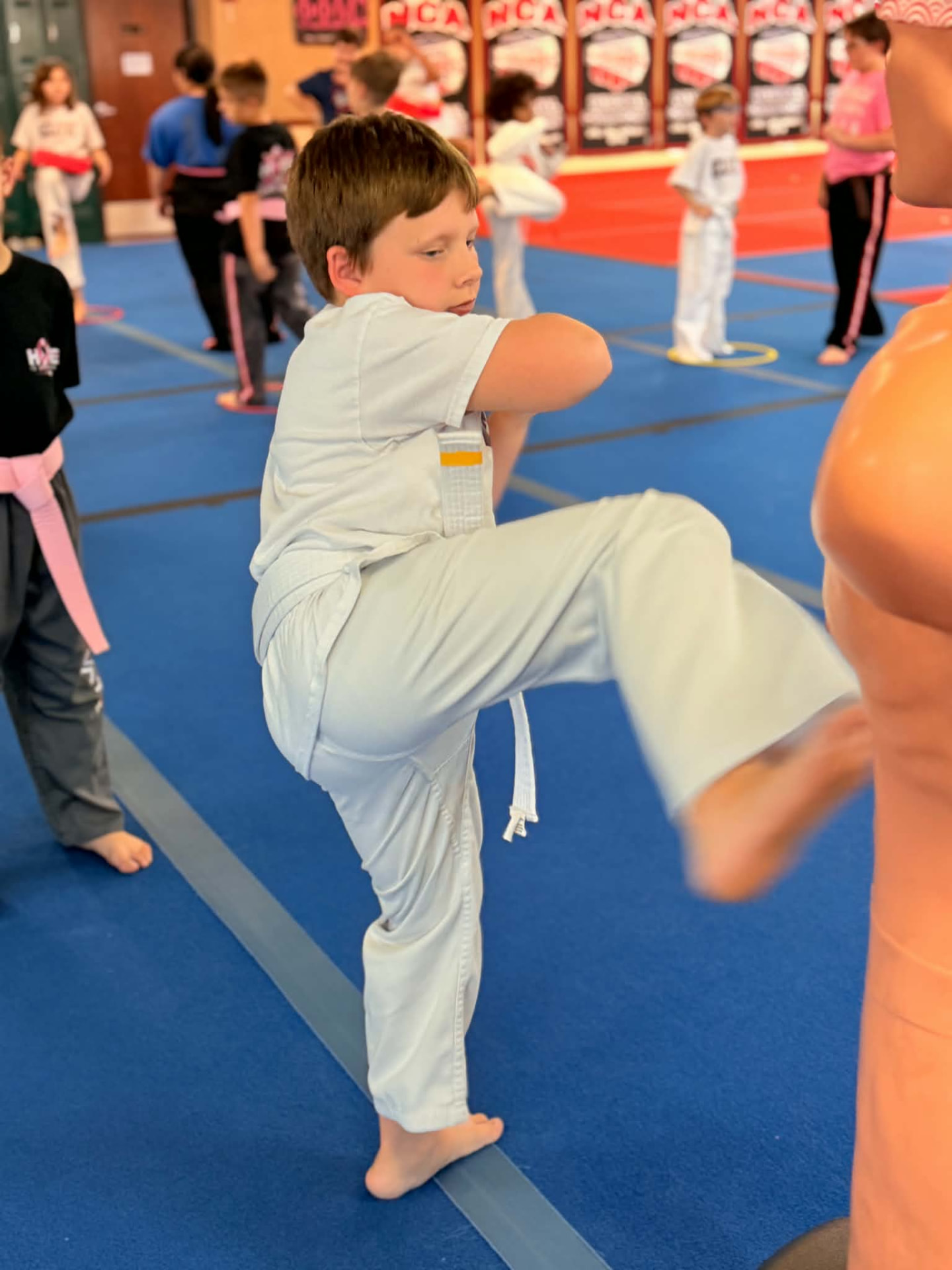 A young Elite Martial Arts athlete in a white gi and white belt practicing a kick against a training dummy on the blue mats.