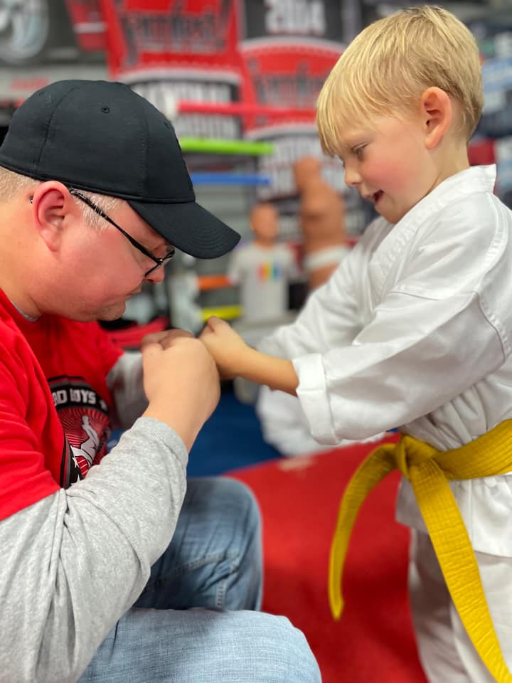 A coach kneeling on the mats guiding a young athlete's hands together as he looks on in his white gi and yellow belt.