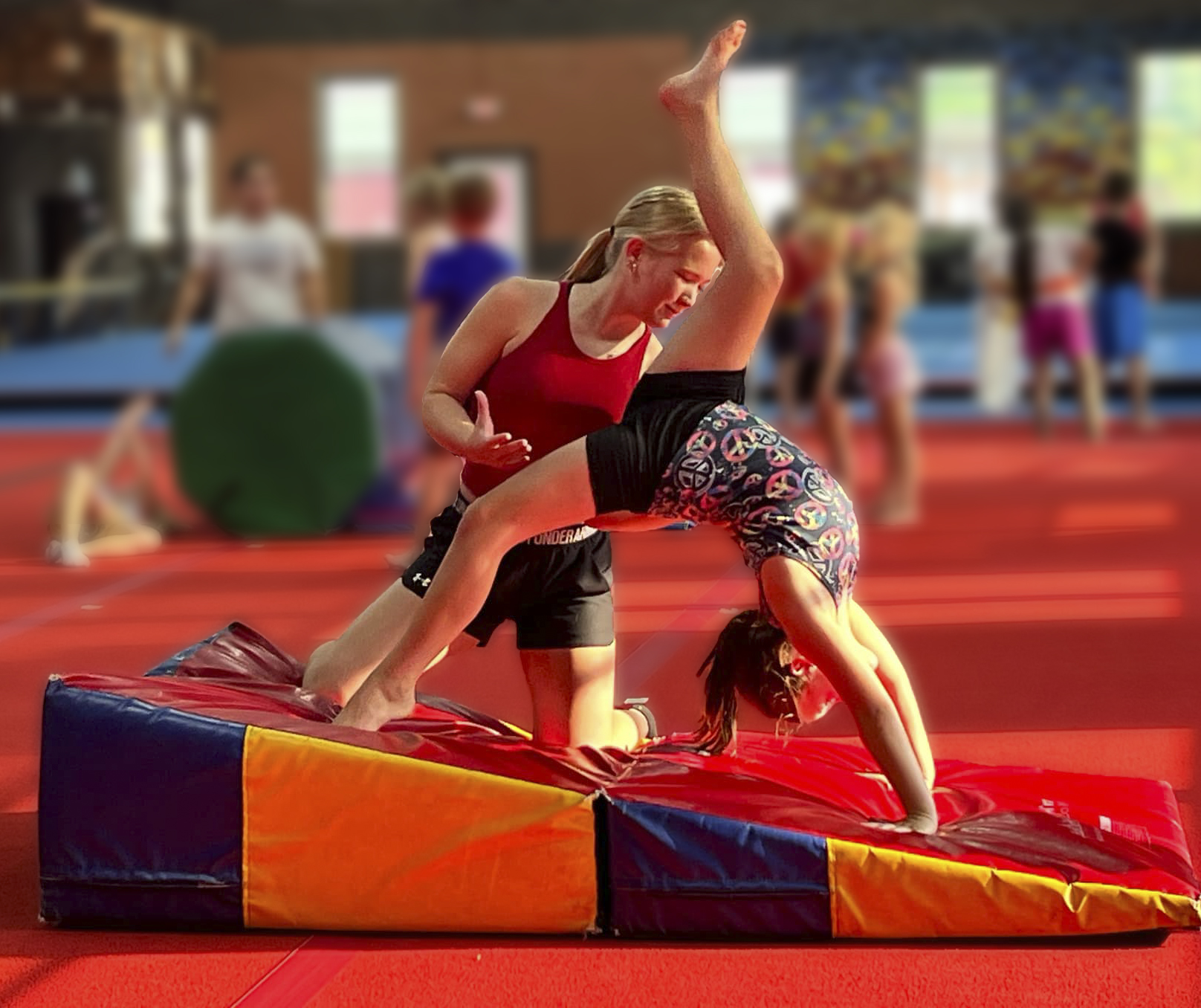 A Pittsburg Elite coach kneeling behind a young athlete, spotting her through a backbend drill on a foam wedge mat.