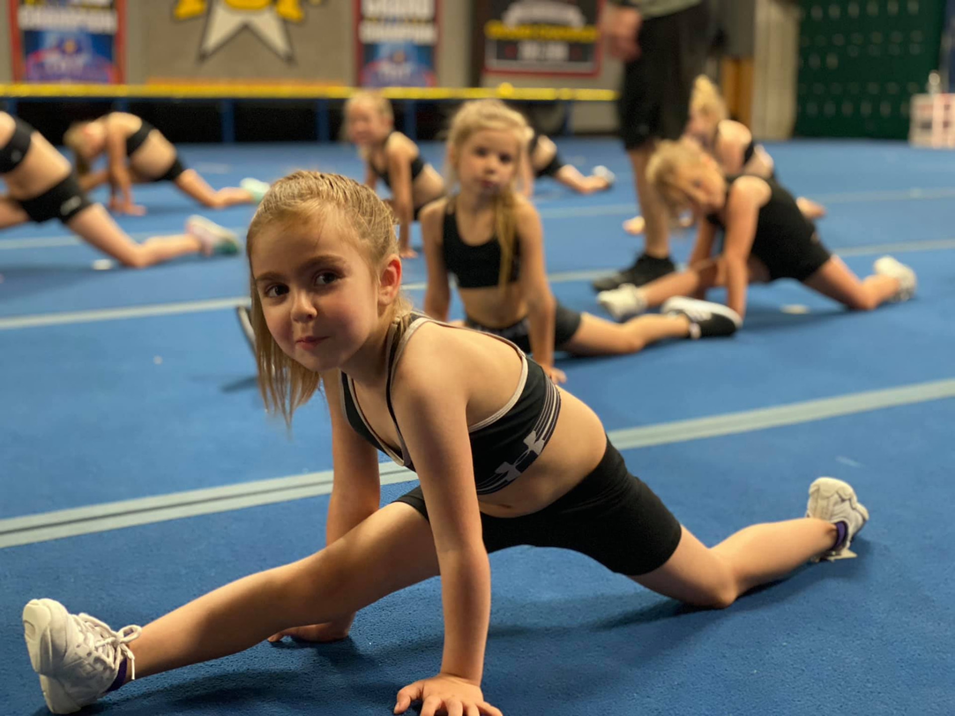 Young athletes working into front splits on the gym floor while a coach stands nearby.